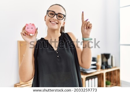Young brunette woman holding piggy bank at the office surprised with an idea or question pointing finger with happy face, number one 