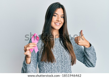 Young brunette woman holding pink cancer ribbon smiling happy and positive, thumb up doing excellent and approval sign 