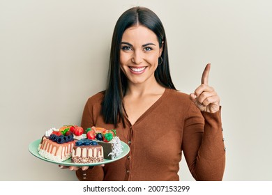 Young brunette woman holding plate with cake slices smiling with an idea or question pointing finger with happy face, number one  - Powered by Shutterstock