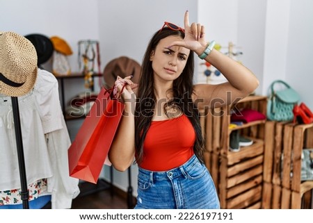 Young brunette woman holding shopping bags at retail shop making fun of people with fingers on forehead doing loser gesture mocking and insulting. 