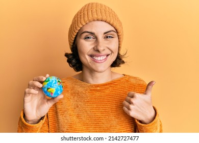 Young brunette woman holding small world ball smiling happy and positive, thumb up doing excellent and approval sign 