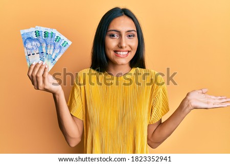 Young brunette woman holding south african 100 rand banknotes celebrating achievement with happy smile and winner expression with raised hand 