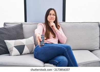 Young brunette woman holding television remote control serious face thinking about question with hand on chin, thoughtful about confusing idea  - Powered by Shutterstock