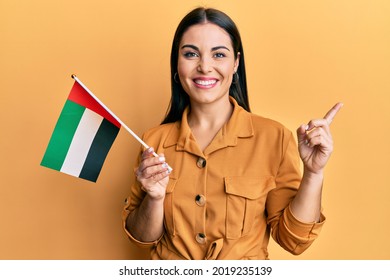 Young brunette woman holding united arab emirates flag smiling happy pointing with hand and finger to the side 