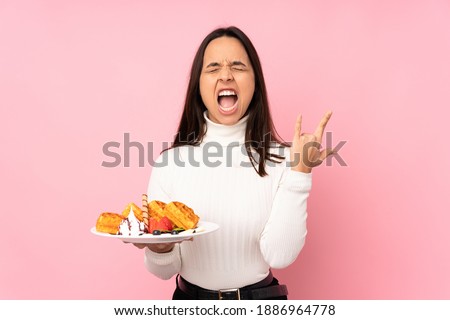 Young brunette woman holding waffles over isolated pink background making rock gesture