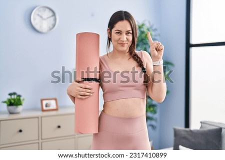 Young brunette woman holding yoga mat smiling with an idea or question pointing finger with happy face, number one 