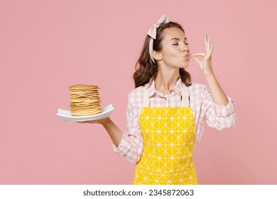 Young brunette woman housewife in yellow apron hold plate with pancakes making okay taste delight sign while doing housework isolated on pastel pink background studio portrait. Housekeeping concept