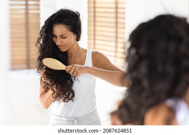 Young Brunette Woman Irritated By Tangled Hair, Using Bamboo Brush In Bathroom, Unhappy Young Female Suffering Painful Feeling, Frowning While Combing Her Curly Hair At Home, Selective Focus