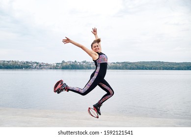 Young brunette woman in kangoo jumps, jumping in the air in front of city lake in summer. Fitness trainer, wearing black fitness overall, doing exercises. Healthy lifestyle concept