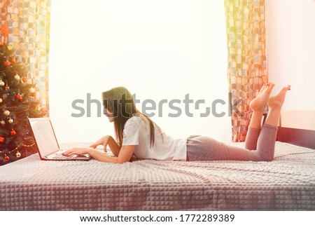 A young brunette woman lies on the bed and uses a laptop. In the background, the interior of the room, a window and a Christmas tree. Side view. Copy space. Sunlight