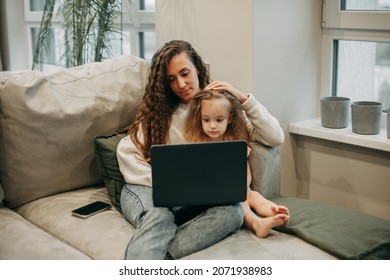 A young brunette woman with long wavy hair in jeans and a white sweater sits at home on the sofa in an embrace with her 2-year-old daughter and watches cartoons behind a laptop, next to a smartphone - Shutterstock ID 2071938983