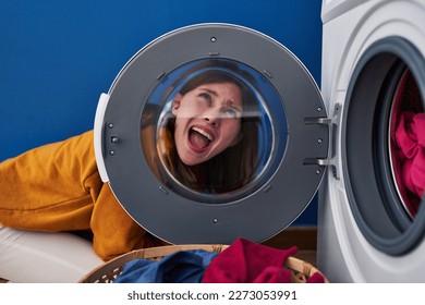 Young brunette woman looking through the washing machine window angry and mad screaming frustrated and furious, shouting with anger looking up. 