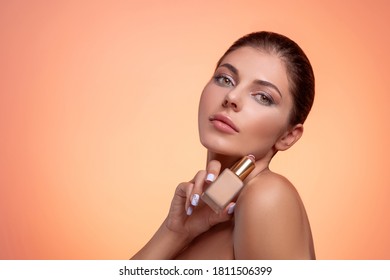 Young Brunette Woman With Naked Shoulders, Natural Makeup And Perfect Clean Fresh Skin Holding Foundation Bottle In Hand. Studio Portrait On Orange Background. Facial Skincare And Anti-age Treatment