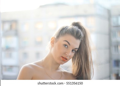 Young Brunette Woman On A Balcony On A Background Of The City. Woman Orange Lips. Portrait Of Beauty Girl. A Girl From A High Ponytail Long Hair