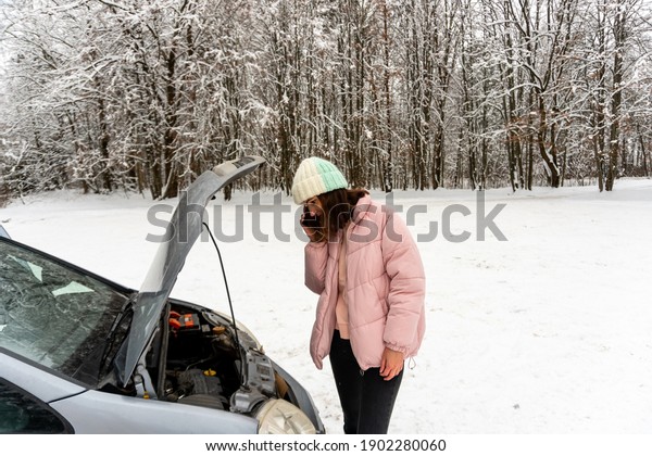 Young brunette woman in a pink winter down jacket
and warm hat is standing near a car with an open hood and talking
on the phone