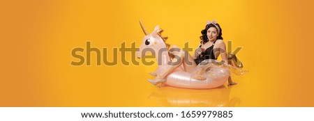 Young brunette woman with pin-up hairstyle in a black swimsuit posing with a beach inflatable unicorn toy on a yellow background