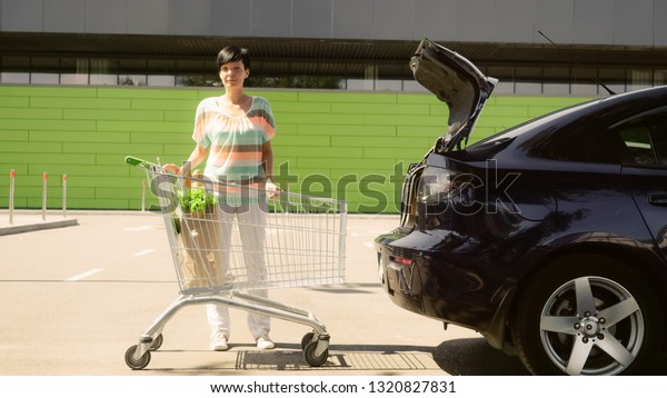 Young brunette woman putting paper bags with\
food and watermelon into car trunk and sits in a car. Girl with\
foodstuffs in paper bag near grocery store. Lady wearing casual\
clothes sunshine\
outdoors.