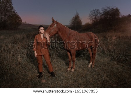 Young brunette woman in a red brown trouser suit posing with a brown horse. Sunset sky and autumn nature. The inscription on the horse: have fun.