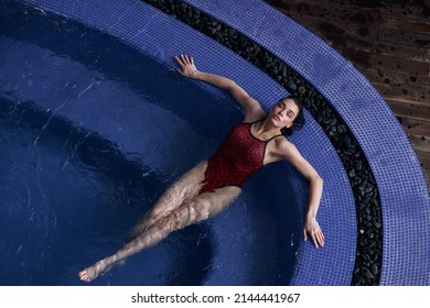 Young Brunette Woman In Red One-piece Swimsuit Rests In Clear Water Leaning On Swimming Pool Edge At Spa Resort View From Above