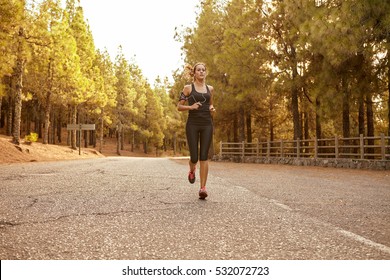 Young Brunette Woman Running In A Forest On A Tarred Road Cutting Through It With Bright Sunshine From Behind Her