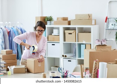 Young brunette woman sealing box with online order of client with cellotape while working in office