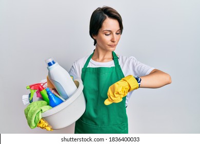 Young Brunette Woman With Short Hair Wearing Apron Holding Cleaning Products Checking The Time On Wrist Watch, Relaxed And Confident 