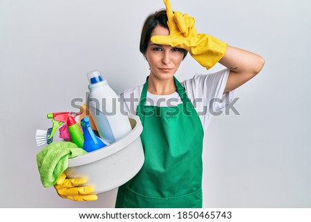 Young brunette woman with short hair wearing apron holding cleaning products making fun of people with fingers on forehead doing loser gesture mocking and insulting. 