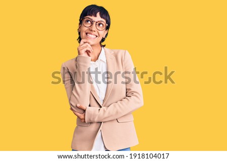 Young brunette woman with short hair wearing business jacket and glasses looking confident at the camera with smile with crossed arms and hand raised on chin. thinking positive. 