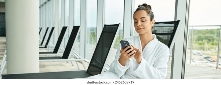 A young brunette woman sitting in a chair, captivated by her cell phone screen, in an indoor spa with a swimming pool. - Powered by Shutterstock