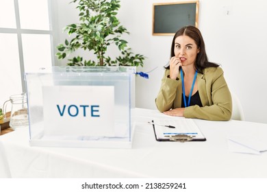 Young Brunette Woman Sitting At Election Table With Voting Ballot Looking Stressed And Nervous With Hands On Mouth Biting Nails. Anxiety Problem. 