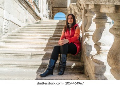 Young brunette woman sitting on a white ladder with balusters. The girl travels to beautiful palaces. Stairs with balusters in the historic courtyard of the building.