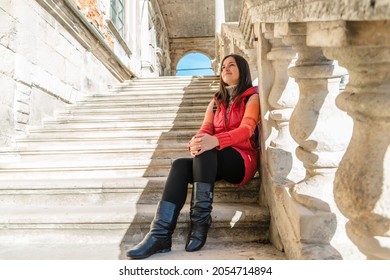 Young brunette woman sitting on a white ladder with balusters. The girl travels to beautiful palaces. Stairs with balusters in the historic courtyard of the building.