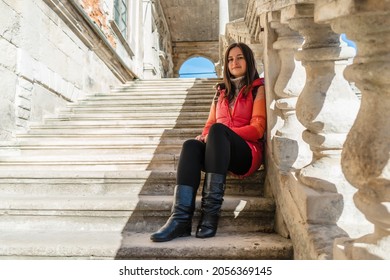 Young brunette woman sitting on a white ladder with balusters. The girl travels to beautiful palaces. Stairs with balusters in the historic courtyard of the building.