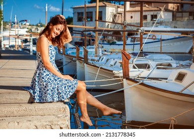 young brunette woman sitting on a dock of Cala Figuera Harbour, wearing a blue and white floral pattern dress 