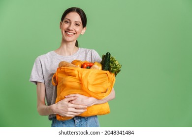 Young brunette woman smiling while holding textile bag with vegetable isolated over green background