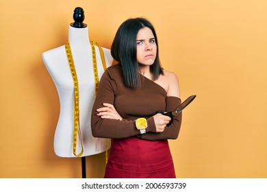 Young Brunette Woman Standing By Manikin Holding Scissors Clueless And Confused Expression. Doubt Concept. 