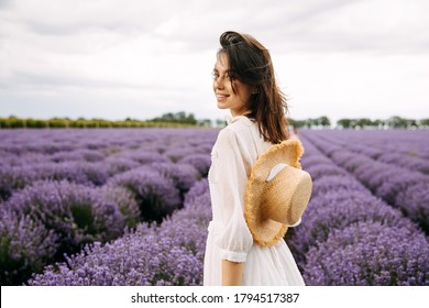 Young brunette woman standing in lavender field, wearing a bohemian white dress and a straw hat.