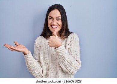 Young brunette woman standing over blue background showing palm hand and doing ok gesture with thumbs up, smiling happy and cheerful 