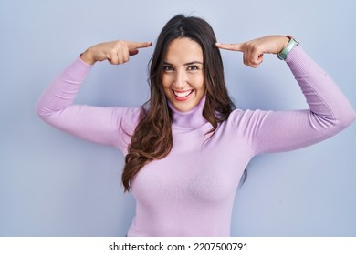 Young Brunette Woman Standing Over Blue Background Smiling Pointing To Head With Both Hands Finger, Great Idea Or Thought, Good Memory 