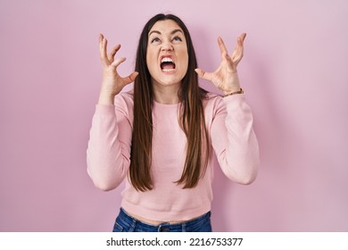 Young brunette woman standing over pink background shouting and yelling with aggressive expression and arms raised. frustration concept.  - Shutterstock ID 2216753377