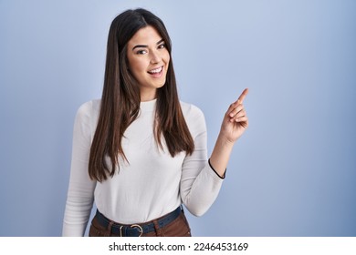 Young brunette woman standing over blue background with a big smile on face, pointing with hand finger to the side looking at the camera.  - Powered by Shutterstock