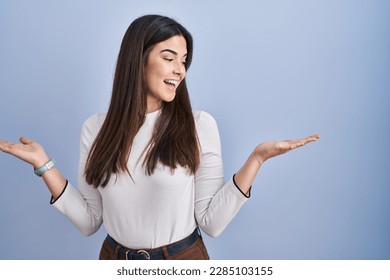 Young brunette woman standing over blue background smiling showing both hands open palms, presenting and advertising comparison and balance  - Powered by Shutterstock