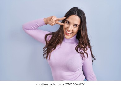 Young brunette woman standing over blue background doing peace symbol with fingers over face, smiling cheerful showing victory 