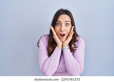 Young brunette woman standing over blue background afraid and shocked, surprise and amazed expression with hands on face  - Powered by Shutterstock