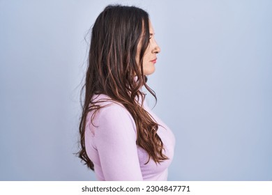 Young brunette woman standing over blue background looking to side, relax profile pose with natural face and confident smile. 