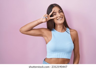 Young brunette woman standing over pink background doing peace symbol with fingers over face, smiling cheerful showing victory  - Powered by Shutterstock