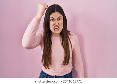 Young brunette woman standing over pink background angry and mad raising fist frustrated and furious while shouting with anger. rage and aggressive concept.  - Shutterstock ID 2341065771
