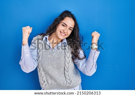 Young brunette woman standing over blue background celebrating surprised and amazed for success with arms raised and eyes closed. winner concept. 