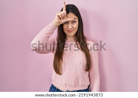 Young brunette woman standing over pink background making fun of people with fingers on forehead doing loser gesture mocking and insulting. 