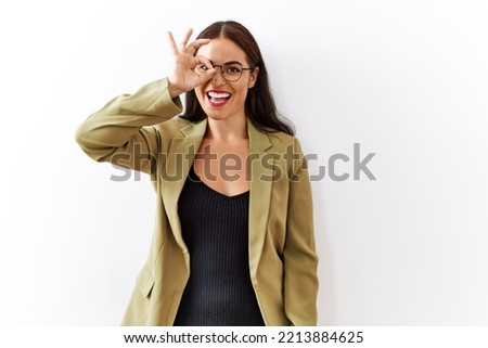 Young brunette woman standing over isolated background doing ok gesture with hand smiling, eye looking through fingers with happy face. 